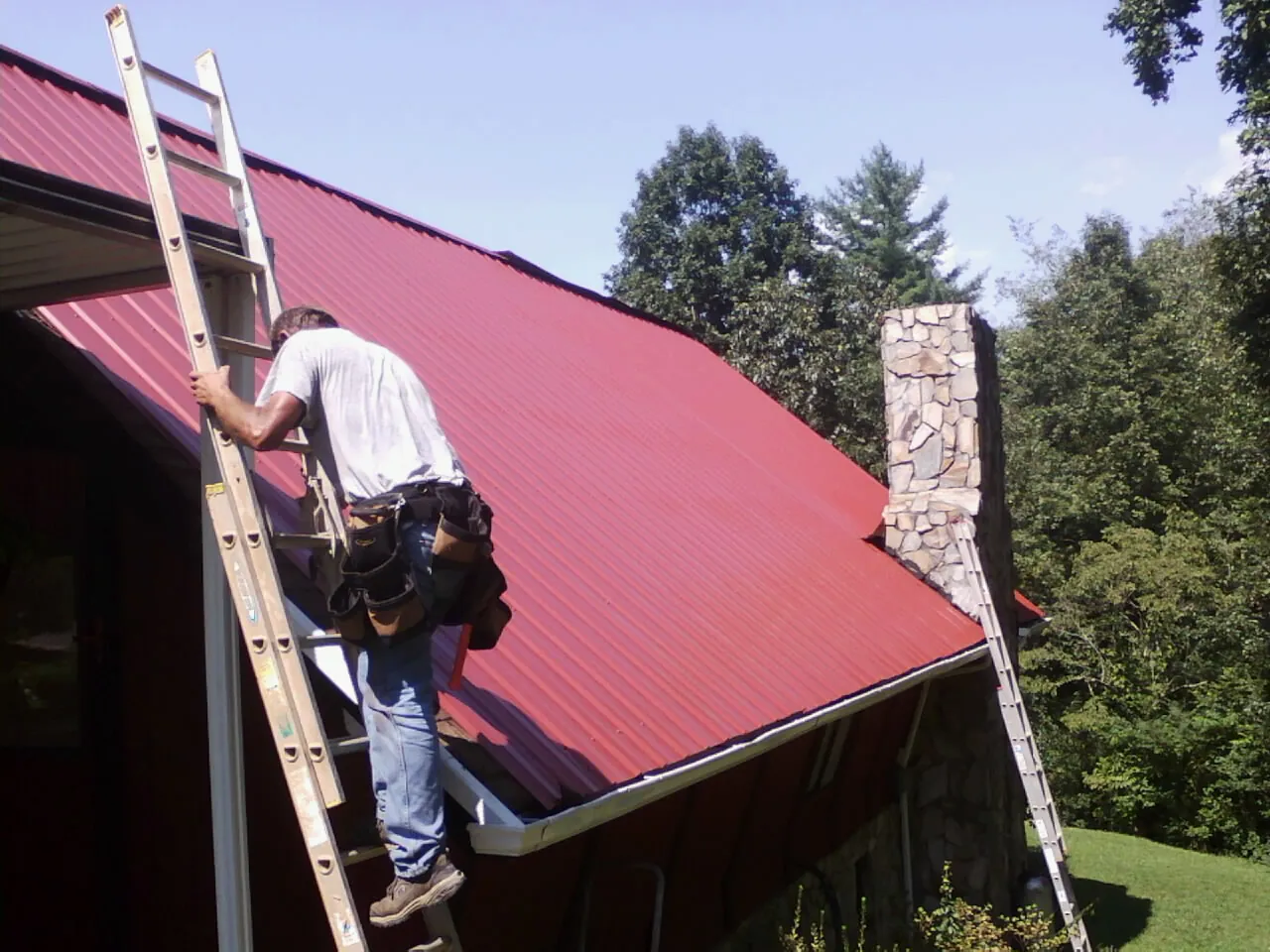 Roofer on a ladder beside a red metal roof and stone chimney — Soffit & Fascia Repair by Roofing Brothers in Harlowton